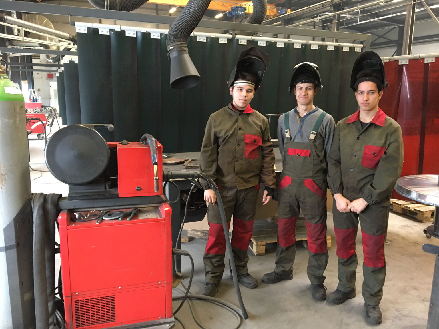 Drei Arbeiter in Schutzkleidung stehen neben einer Schweißmaschine in einer Werkstatt.