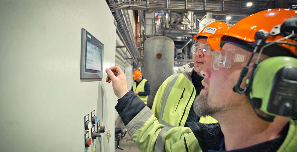 Two workers in protective gear are operating a control panel at an industrial facility.